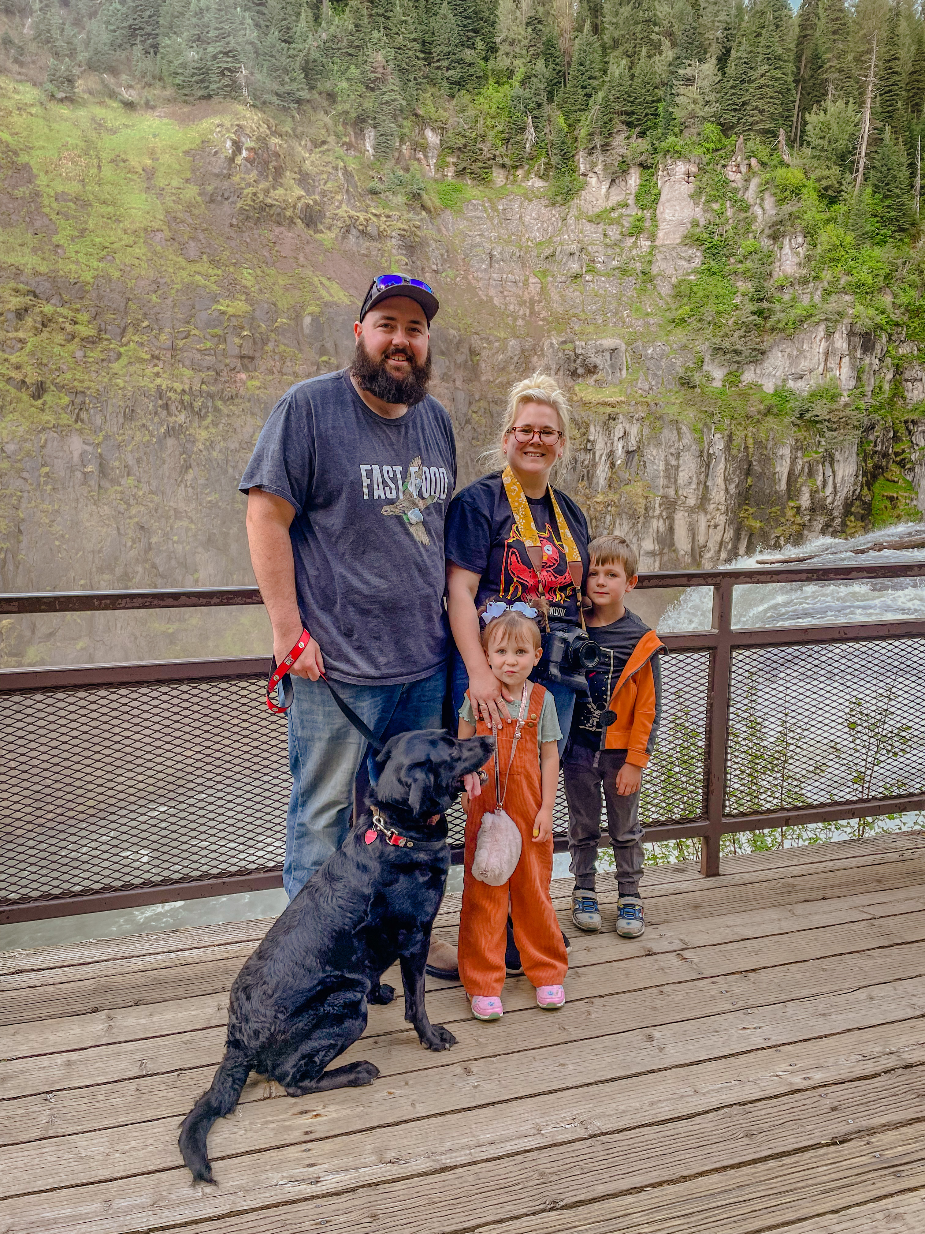 large bearded man, blonde woman, two small children and a dog stand in front of a water fall.